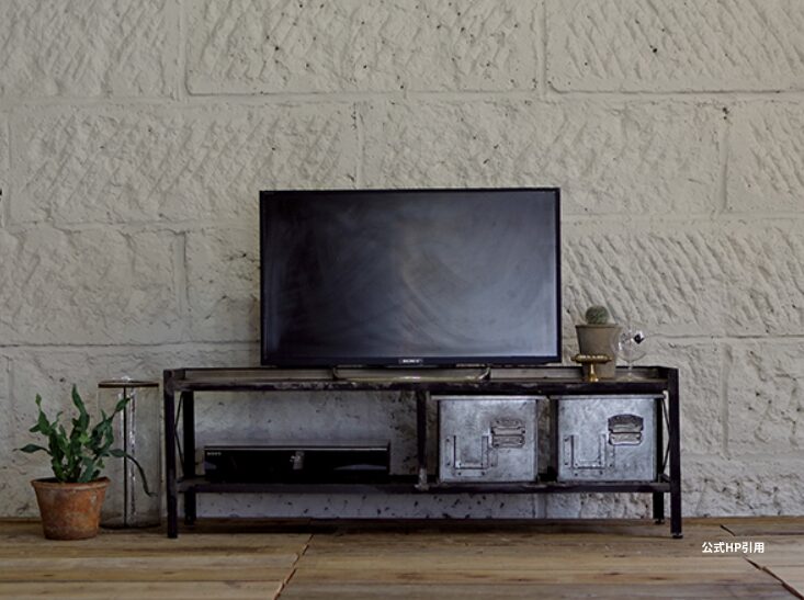 インテリアコーディネート画像：アイアンフレームのTVボードにテレビと収納ボックスを組み合わせ、無骨なレンガ調壁と木製フロアでまとめたインダストリアルテイストの落ち着いたリビング空間