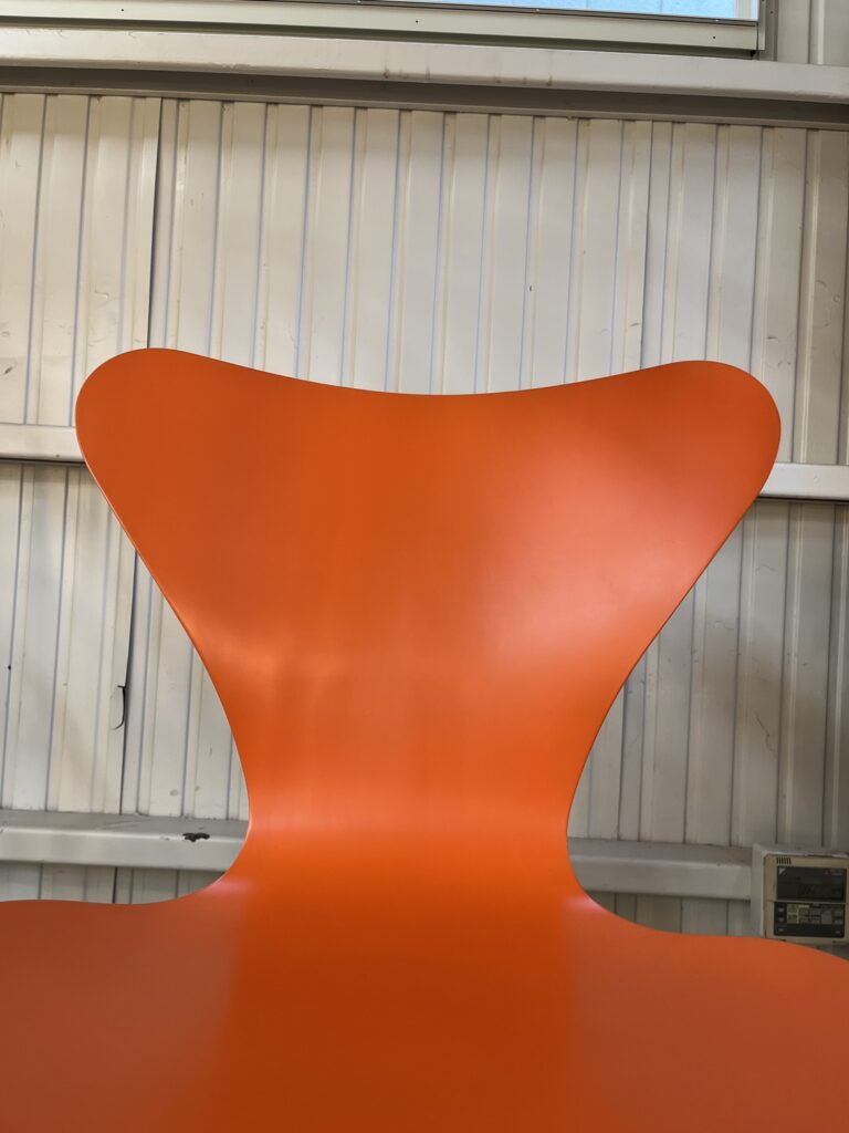FRITZ HANSEN(フリッツ・ハンセン)のセブンチェア。木目を見せないオレンジのラッカー仕上げが特徴で、滑らかなシェルの曲線と細いスチールパイプ脚が美しく調和するデザイン。